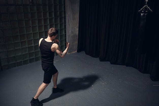 Top rear view of athletic fir young man with muscular arms wearing black clothes while boxing, punching invisible enemy, standing isolated in dark room, casting shadow on gray concrete floor