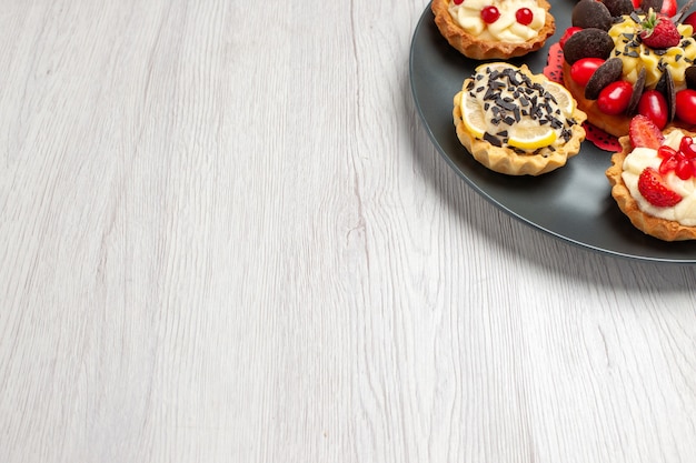 Top right side view chocolate cake rounded with berry tarts in the grey plate at the top right of the white wooden table