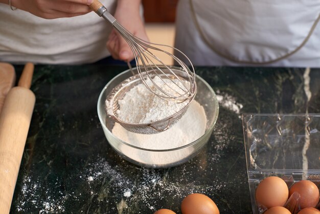 Top shot of unrecognizable people in aprons sieving flour into bowl, and eggs on table
