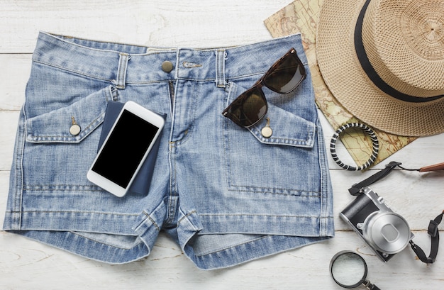 Top view accessoires to travel with women clothing concept.White mobile phone,hat,map,camera,necklace,trousers and sunglasses on white wood table.