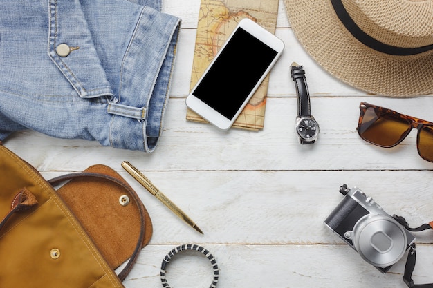 Вид сверху, чтобы путешествовать с концепцией женской одежды. Белый мобильный телефон, часы, сумка, шляпа, карта, камера, ожерелье, брюки и солнцезащитные очки на белом деревянном столе.