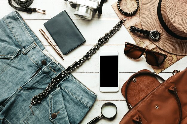 top view accessoires to travel with women clothing concept.white mobilephone, watch,bag,hat,map,camera,necklace,trousers and sunglasses on white wood table.