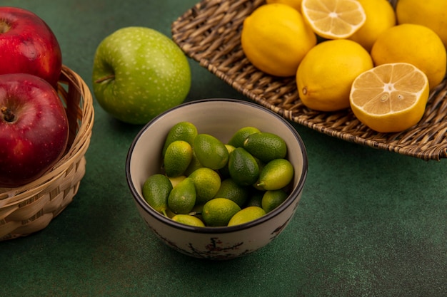 Вид сверху лимонов со вкусом кислоты на плетеном подносе с кинканами на миске с красными сладкими яблоками на ведре на зеленой поверхности