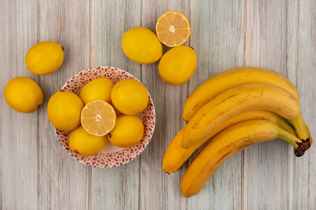 Вид сверху на цельные лимоны со вкусом кислоты на миске с бананами, изолированными на сером деревянном фоне