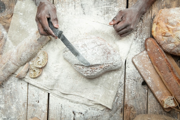 Вид сверху афро-американский мужчина готовит свежие хлопья, хлеб, отруби на деревянном столе. Вкусная еда, питание, крафтовый продукт. Безглютеновая пища, здоровый образ жизни, экологически чистое и безопасное производство. Ручной работы.