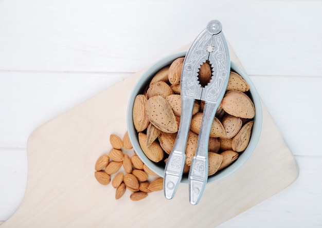 Top view of almond in a bowl with nut cracker on wooden board on white background