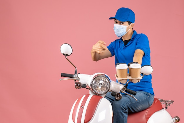 Top view of ambitious courier man in mask wearing hat sitting on scooter showing orders on pastel peach background