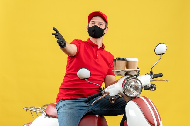 Top view of ambitious delivery guy wearing uniform and hat gloves in medical mask sitting on scooter showing orders speaking about something