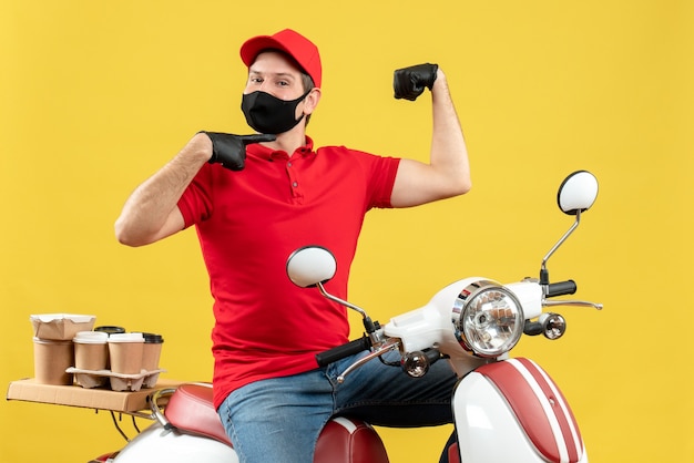 Top view of ambitious young adult wearing red blouse and hat gloves in medical mask delivering order sitting on scooter showing his muscular