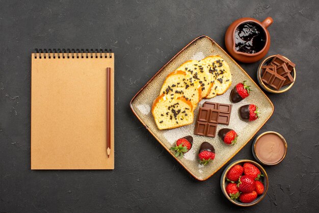 Top view appetizing cake cake with strawberries and chocolate between bowls of chocolate cream strawberries and chocolate next to the brown pencil and cream notebook