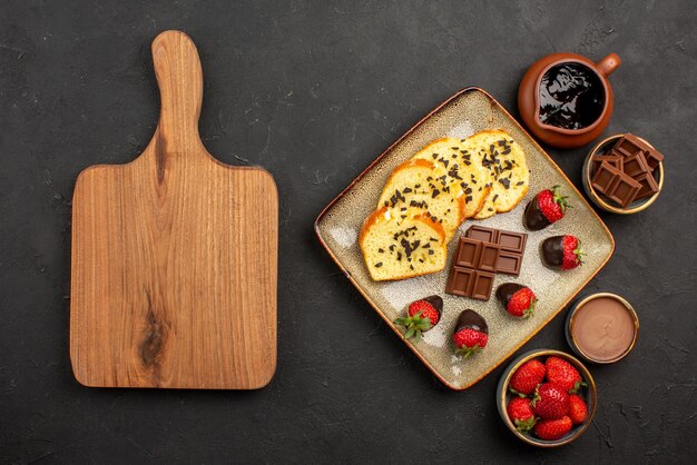 Top view appetizing cake cake with strawberries and chocolate between bowls of chocolate cream strawberries and chocolate next to the brown wooden cutting board