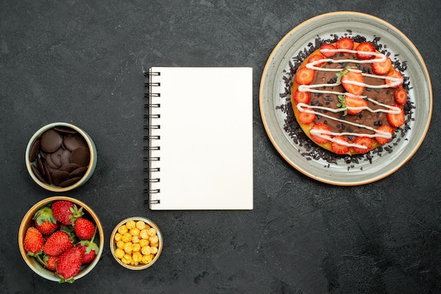 Top view appetizing cake white notebook between cake with chocolate and strawberry pieces and bowls of chocolate strawberry and hazelnut on black table