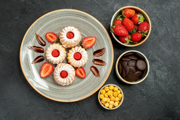 Top view appetizing cookies with sweets bowls of hizelnuts strawberry and chocolate next to cookies with chocolate and strawberry on dark table