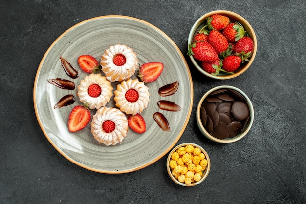 暗いテーブルの上のチョコレートとイチゴのクッキーの横にあるハイゼルナッツイチゴとチョコレートのスイーツボウルと食欲をそそるクッキーの上面図