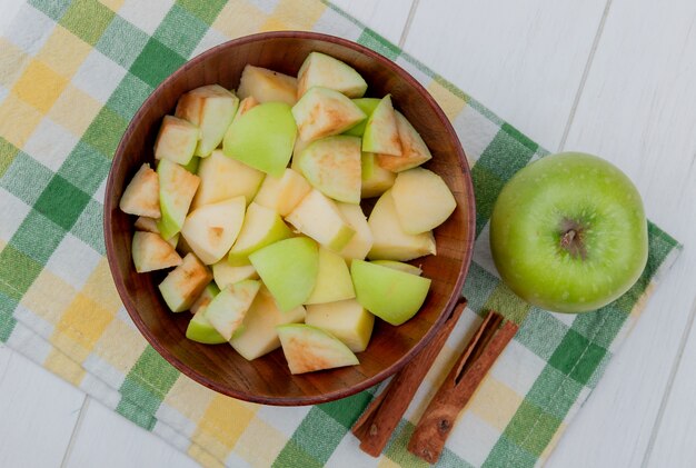 top view of apple cubes in bowl and whole one with cinnamon on plaid cloth and wooden