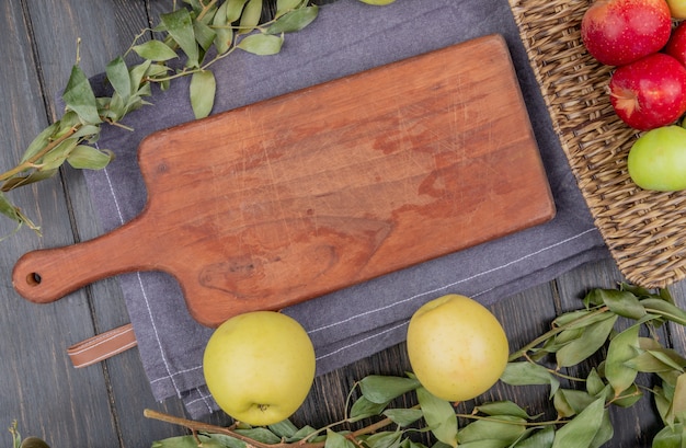 Top view of apples around cutting board on gray cloth with leaves on wooden background