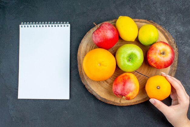 Top view apples lemon oranges on wood board notebook orange in woman hand on dark surface