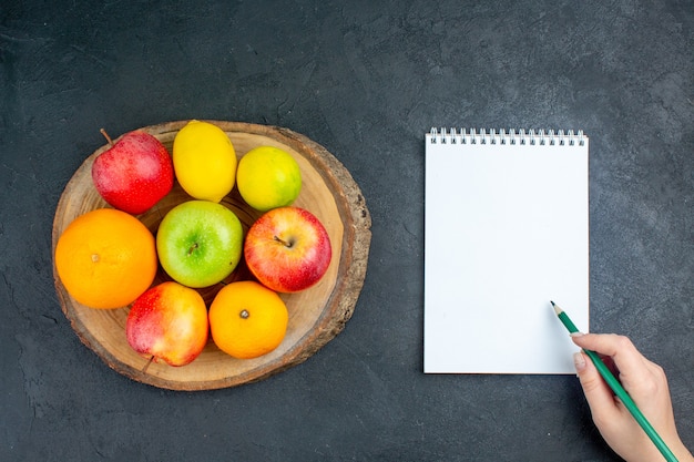 Top view apples lemon oranges on wood board notebook pencil in female hand on dark surface