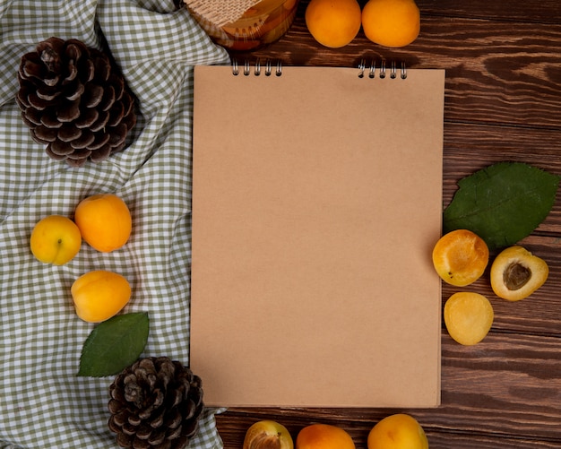 Top view of apricots around note pad with pinecones on wooden background decorated with leaves with copy space