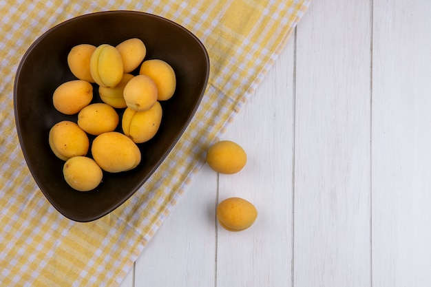 Top view of apricots in a bowl on a checkered yellow towel on a white surface