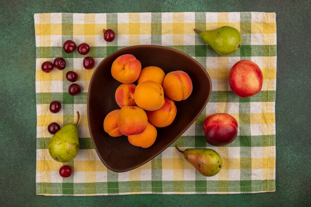 Top view of apricots in bowl and pattern of pear peach cherry on plaid cloth on green background