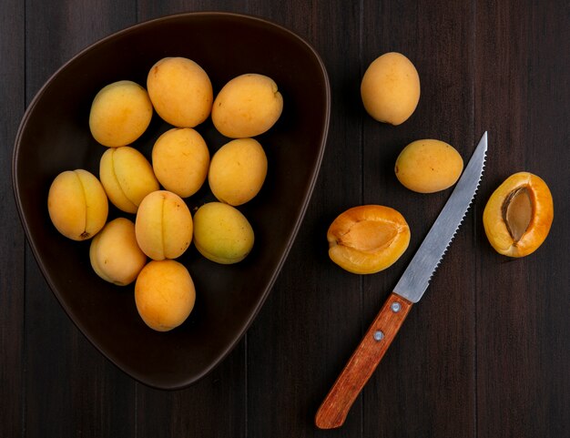 Top view of apricots in a bowl with a knife on a wooden surface