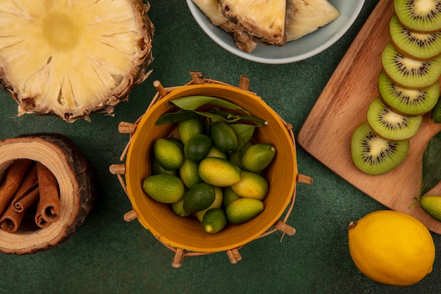 緑の背景に分離されたレモンとシナモンスティックとボウルにパイナップルとパイナップルと木製のキッチンボードにキウイスライスとバケツの芳香族キンカンの上面図