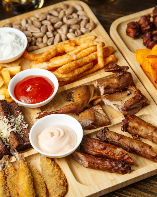 Top view of assorted beer snacks as roasted quails  french fries   pistachios and  potato chips with sauces on a wooden board