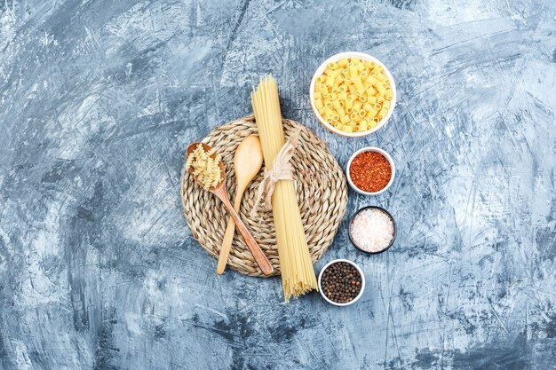 Top view assorted pasta in bowl and wooden spoon with spices on grey plaster and wicker placemat background. horizontal