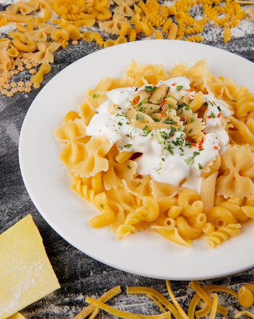 Top view of assorted raw italian pasta and cooked farfalle pasta with cream sauce in a white plate on black background