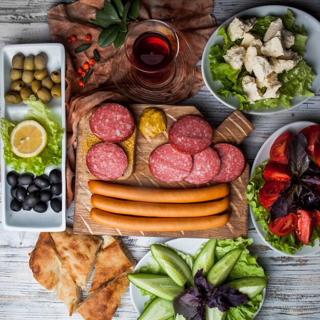 Top view assorted sausages with assorted olives and vegetables and glass of tea in board cookware