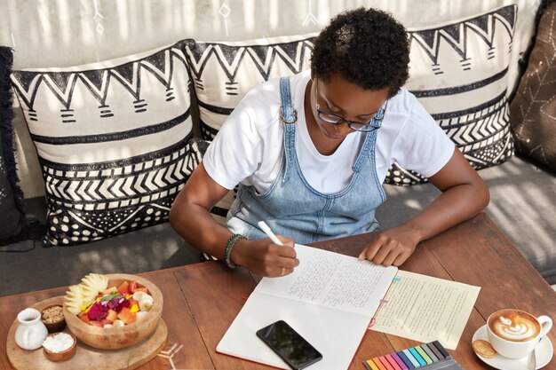 Top view of attractive stylish black woman writes review of book in notepad