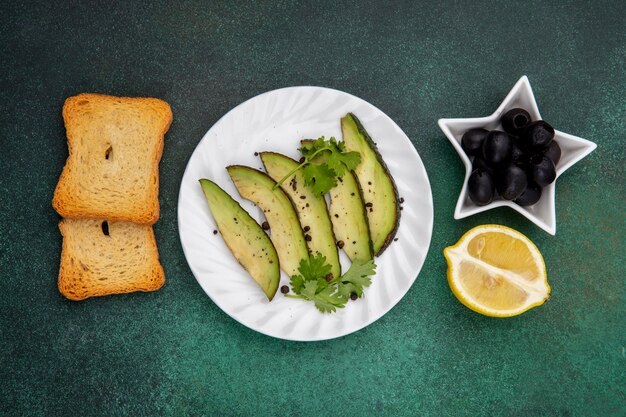 Top view of avocado slices on white plate with black olives a toasted slices of bread and lemon on gre