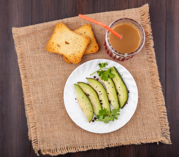 Top view of avocado slices on white plate with toasted slice of bread with juice in a glass on sack cloth on wood