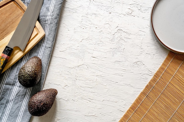 Top view of avocados on a white surface, with a cutting board, knife, and a plate, copy space