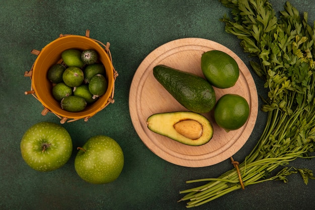 Free photo top view of avocados on a wooden kitchen board with limes with feijoas on a bucket with green apples and parsley isolated on a green wall