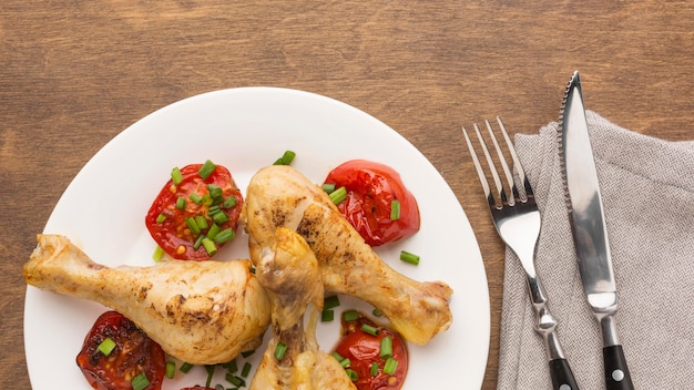 Top view baked chicken drumsticks and tomatoes on plate with cutlery and kitchen towel
