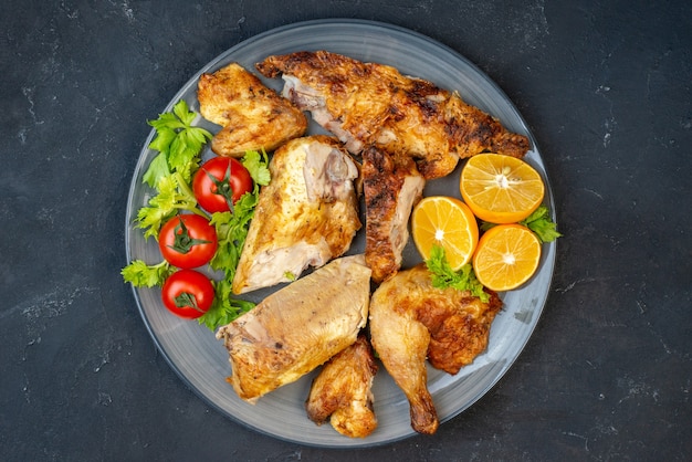 Вид сверху запеченный цыпленок, свежие помидоры, ломтики лимона на круглой тарелке на черном столе