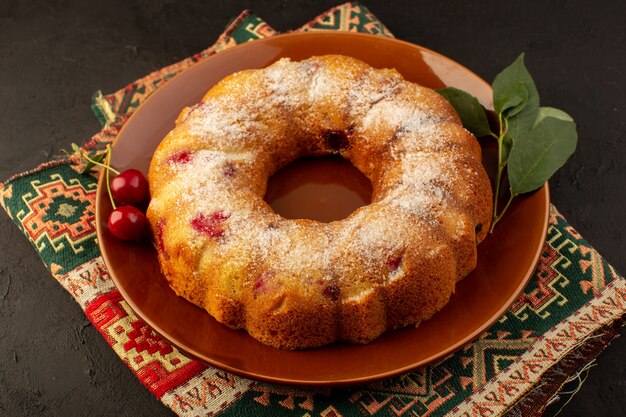 A top view baked fruit cake delicious round with red cherries inside and sugar powder inside round brown plate on dark