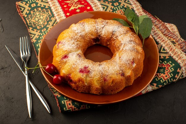 A top view baked fruit cake delicious round with red cherries inside and sugar powder inside round brown plate on dark