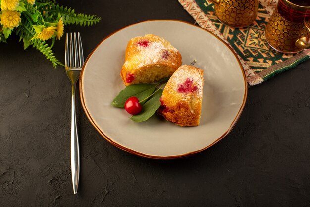 A top view baked fruit cake delicious slices with red cherries inside and sugar powder inside round grey plate on dark