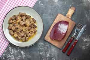 Free photo top view baked liver and onion on oval plate on tablecloth raw liver on cutting board knife and fork