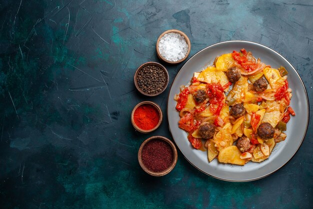 Top view baked potatoes with meatballs and vegetables along with seasonings on dark-blue desk.