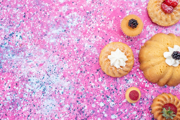 Вид сверху запеченных вкусных пирожных со сливками вместе с ягодами на ярко-фиолетовом столе, бисквитный ягодный сладкий чай для выпечки