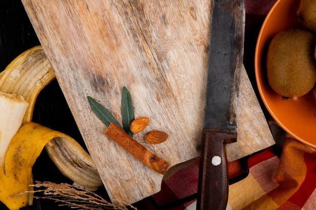 Вид сверху банановых фруктов с миндалем, палочки корицы и старый кухонный нож на деревянной разделочной доске на черном
