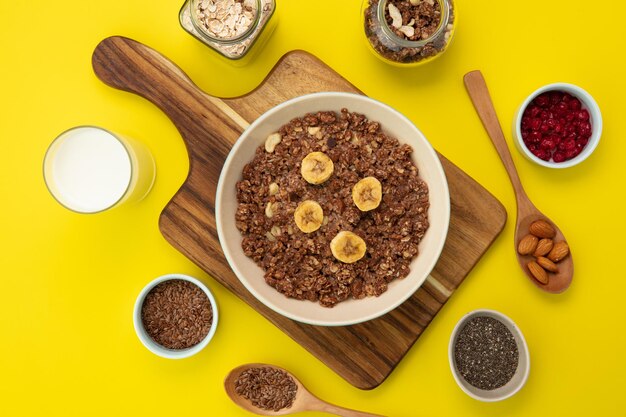 Top view of banana walnut oatmeal in bowl on cutting board with milk flaxseeds oat red currant chia seeds spoonful of almond on yellow background