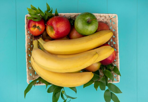 ターコイズブルーの表面に色リンゴと桃の枝が付いているバスケットの桃とバナナのトップビュー