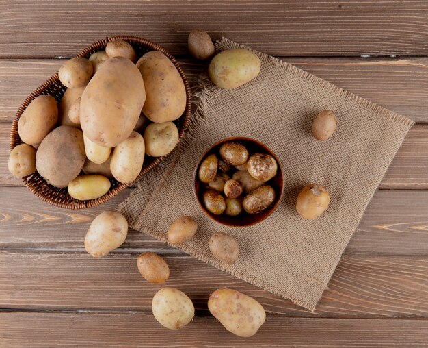 Top view of basket and bowl full of potato on sackcloth on wooden background with copy space