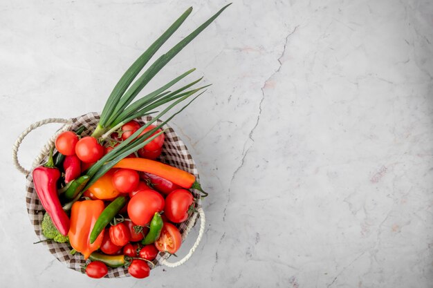 Top view of basket full of vegetables as scallions tomatoes peppers on left side on white surface