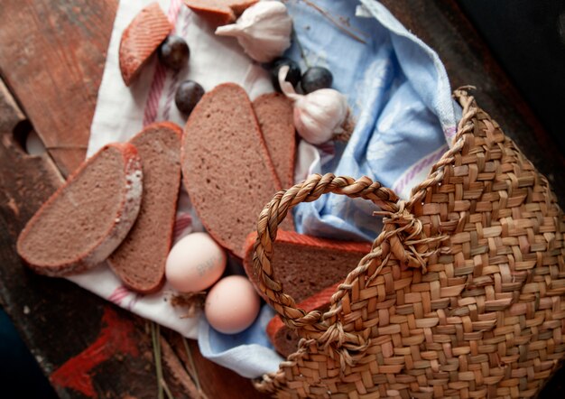 Вид сверху корзина с хлебом ломтиками яиц, сливы и чеснока вокруг на деревянном столе.